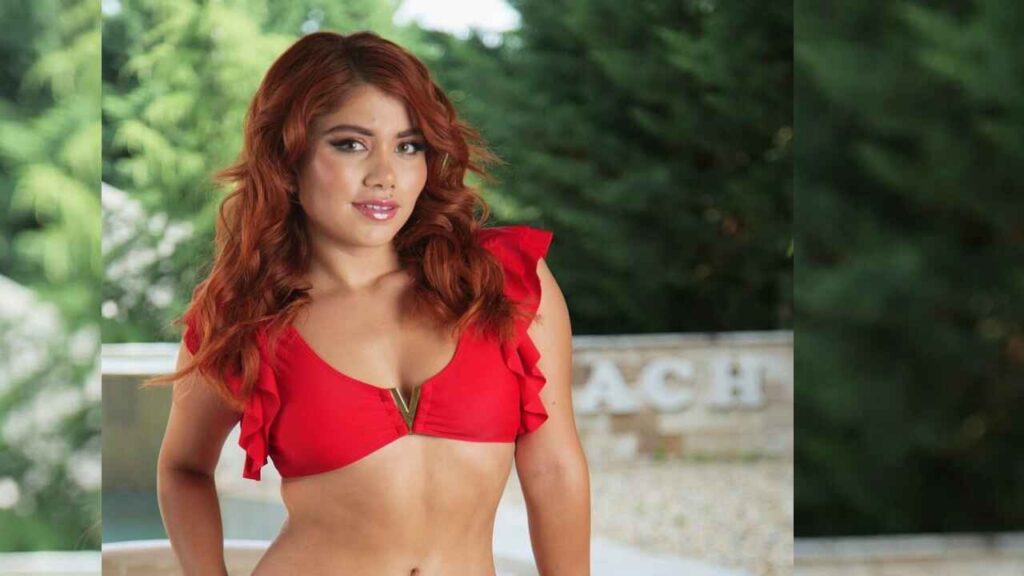 Marina Gold wearing a red ruffle-shoulder bikini top stands outdoors, with green trees and a blurred wooden fence behind her.