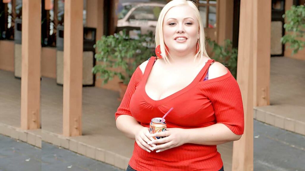 Bunny DeLa Cruz in a red top holds a soda can, standing in an outdoor area with plants and walls made of wood.