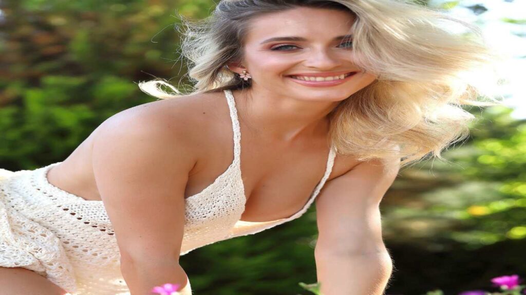 Cara Mell in a white crochet outfit leans forward among colorful flowers, surrounded by lush greenery and sunlight.