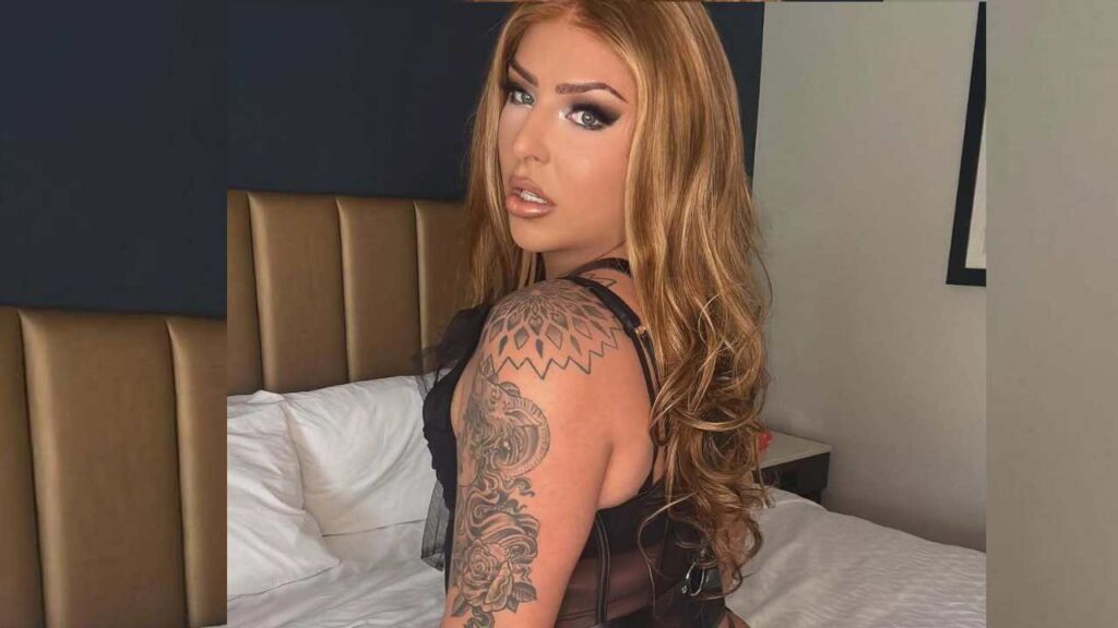 Chanel Santini with long, wavy hair poses in a black outfit, showcasing a detailed tattoo on her shoulder against a hotel room backdrop.