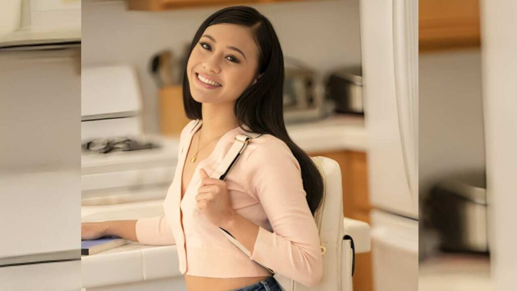 Jasmine Grey wearing a light pink cardigan and jeans, standing in a kitchen with modern appliances and wooden cabinetry.