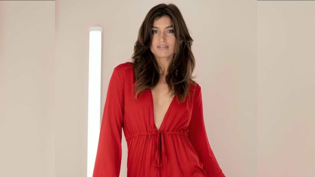 Julia Roca in a deep V-neck red dress stands against a neutral backdrop, exuding elegance and style. Soft lighting enhances the scene.