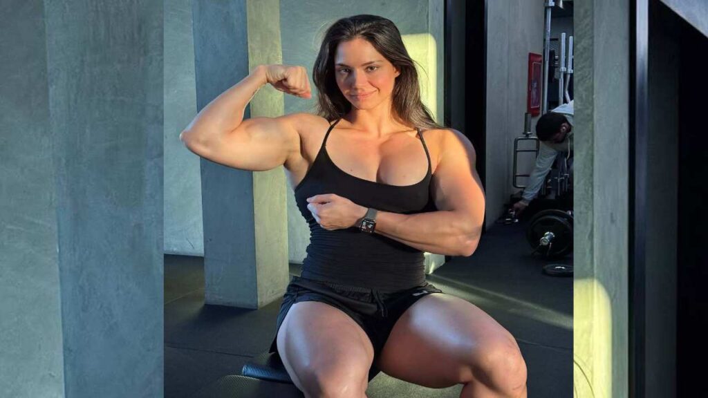 Vladislava Galagan flexes her arm while seated in a gym, showcasing her defined biceps and strong physique.