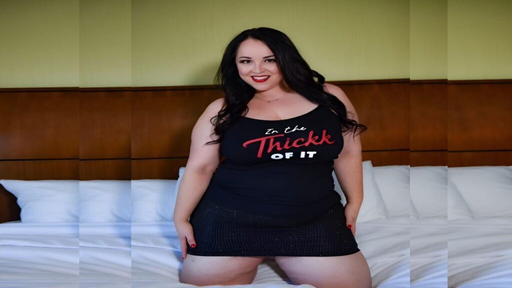 Amanda Thickk in a black tank top and skirt poses confidently on a bed, showcasing a stylish and relaxed atmosphere.