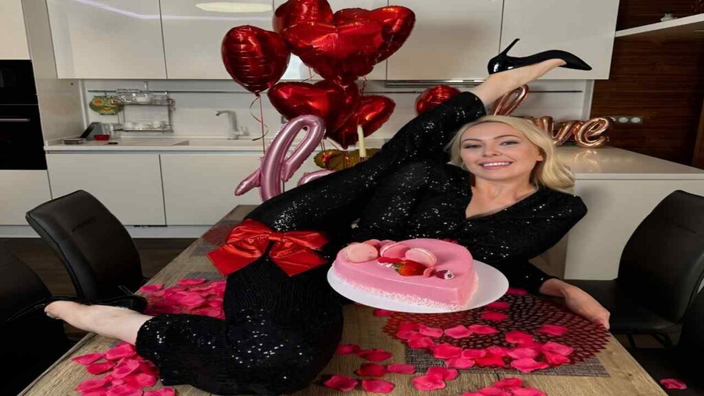 Anna Delos in a black sequin dress sits on a table beside a decorated cake, smiling at the camera