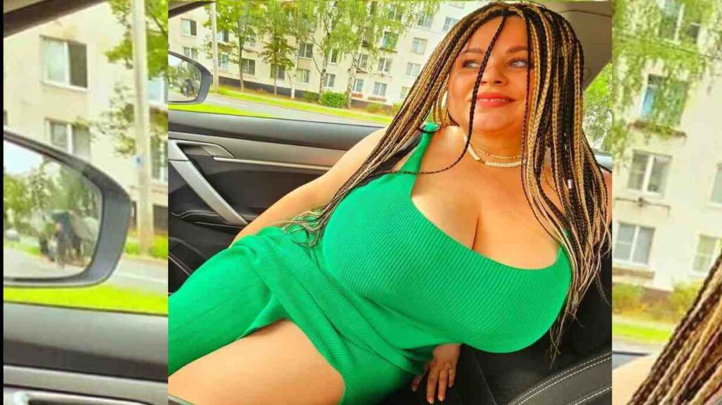 Anna Katz in a green dress with long braided hair sits in a car, with a view of urban buildings and trees in the background.