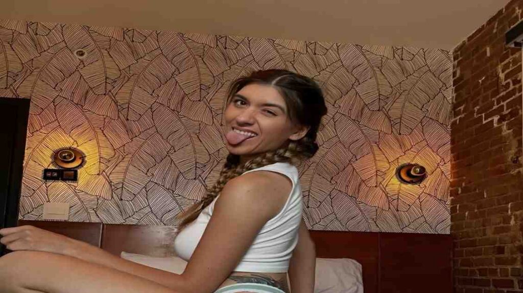 Aubry Babcock sitting on a bed playfully sticking her tongue out, smiling at the camera.