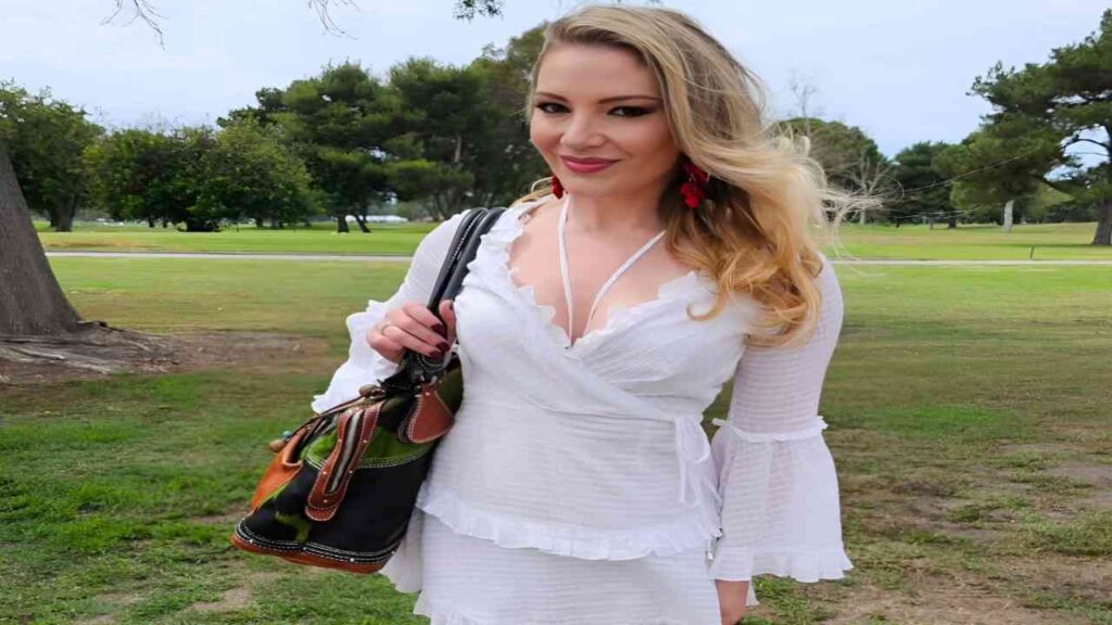 Aurora Snow in a white dress and red heels holds a purse, standing confidently with a stylish pose