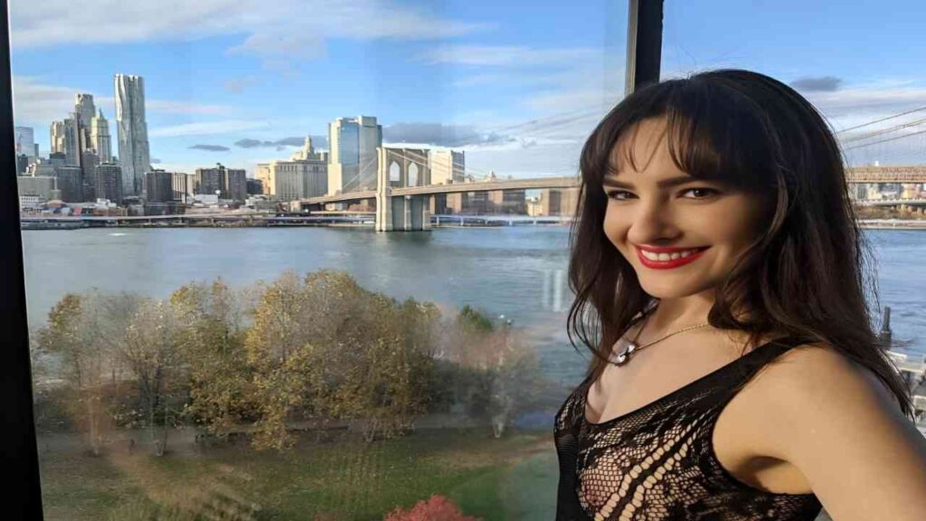 Ava Alex poses for a photo by a window, showcasing a city skyline view in the background