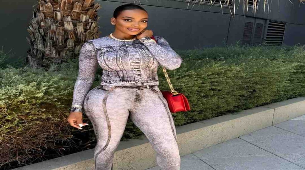 Bailey Brooke wearing a grey jumpsuit holds a red bag, standing confidently against a neutral background