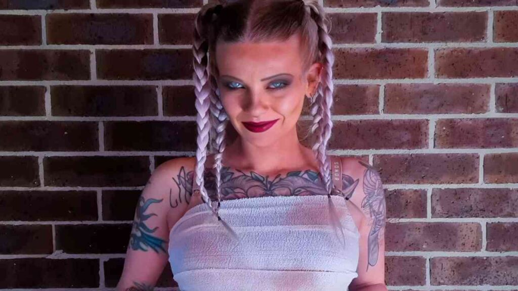 Coral Jade with tattoos and braided hair stands confidently in front of a textured brick wall