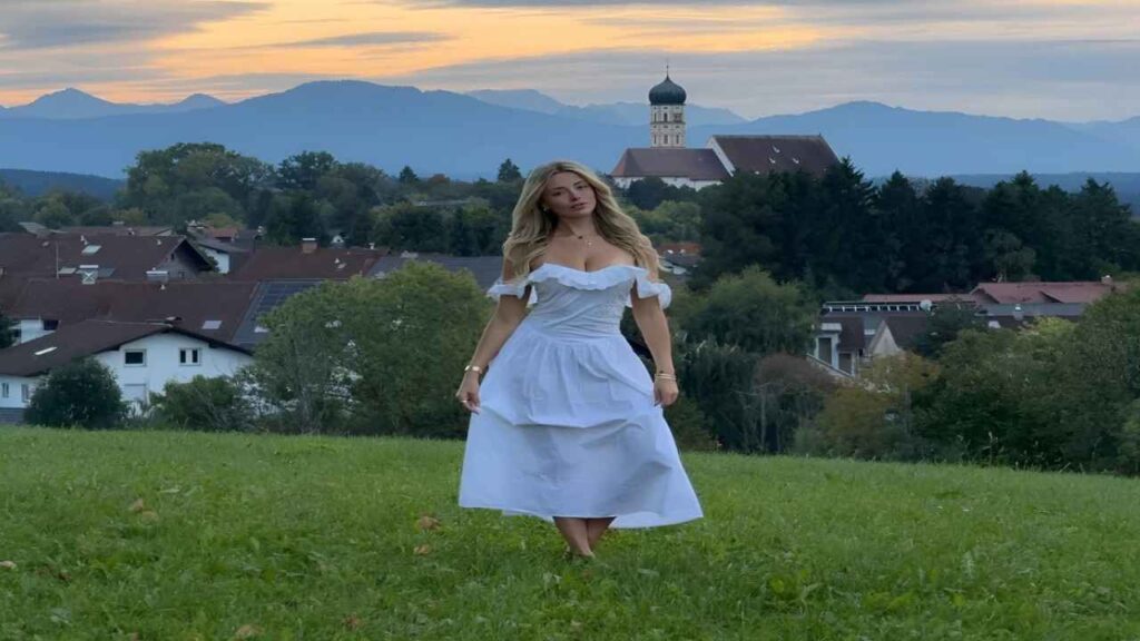 Corinna Kopf in a flowing white dress stands gracefully in a sunlit field, surrounded by tall grass and wildflowers