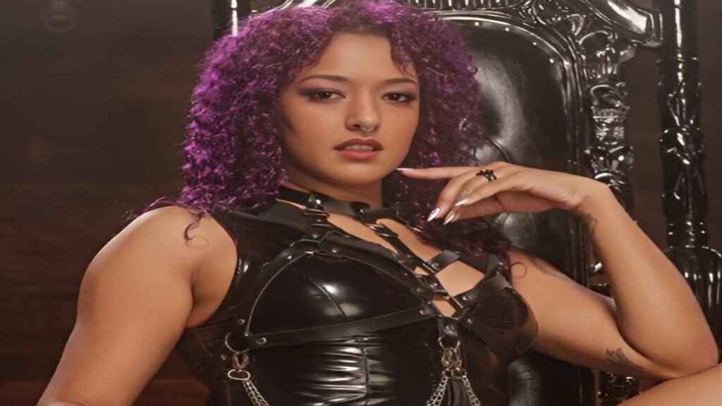 Daisy Ducati with purple hair dressed in leather sits confidently on a chair, exuding a bold and stylish presence