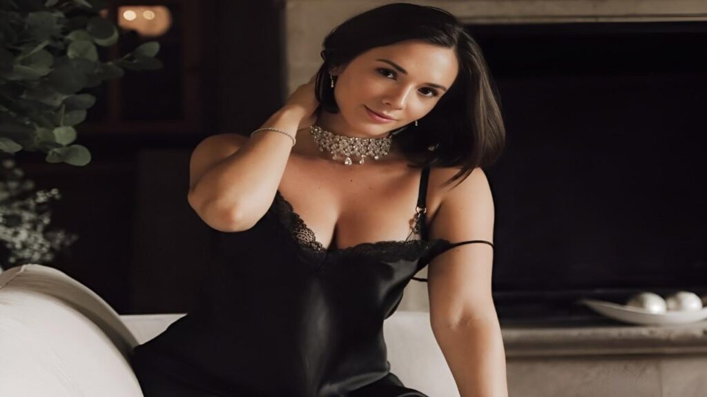 Eve Sweet in a black dress is seated on a couch, looking thoughtfully to the side.