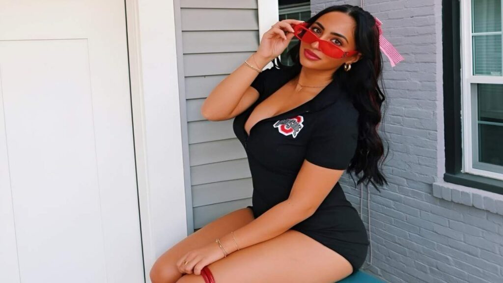 Giana Taylor wearing a short black dress and red sunglasses poses confidently against a neutral background.