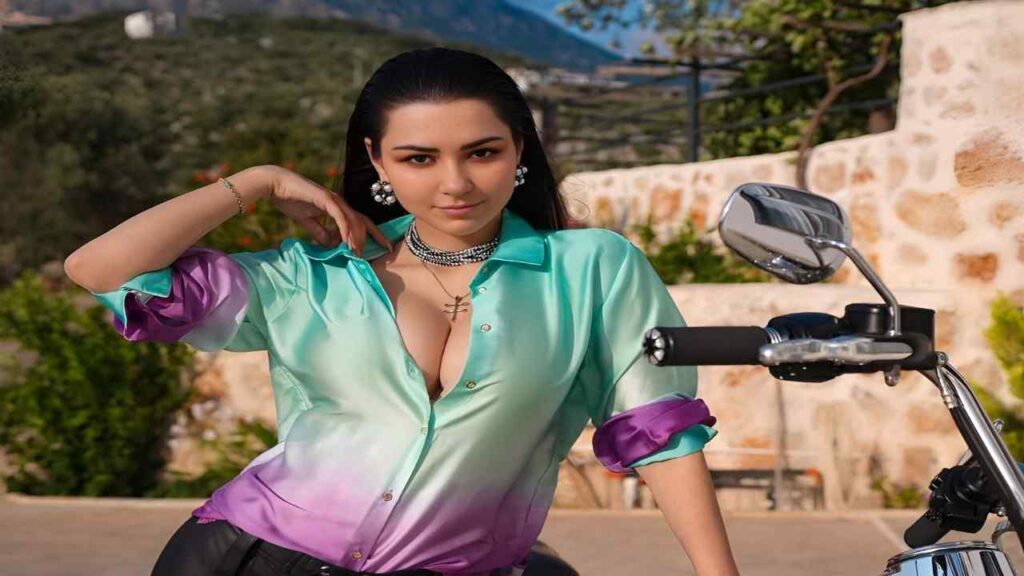 Helga Lovekaty wearing a purple shirt and green pants sits confidently on a motorcycle.