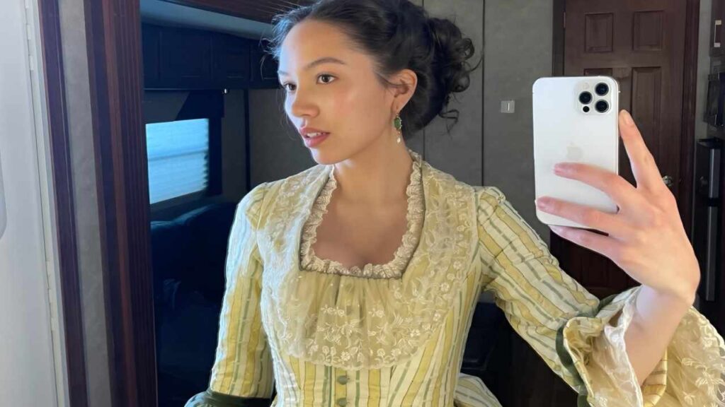 Jasmine Vega in a yellow dress smiles as she takes a selfie with her phone