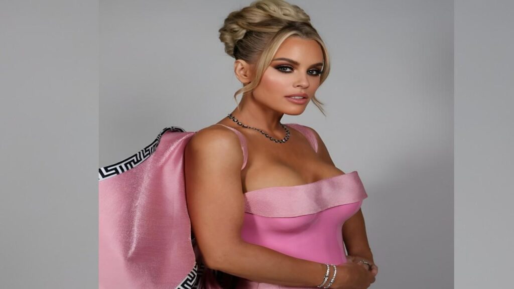 Jenny McCarthy in a pink dress poses for a photo, smiling and standing confidently against a neutral background