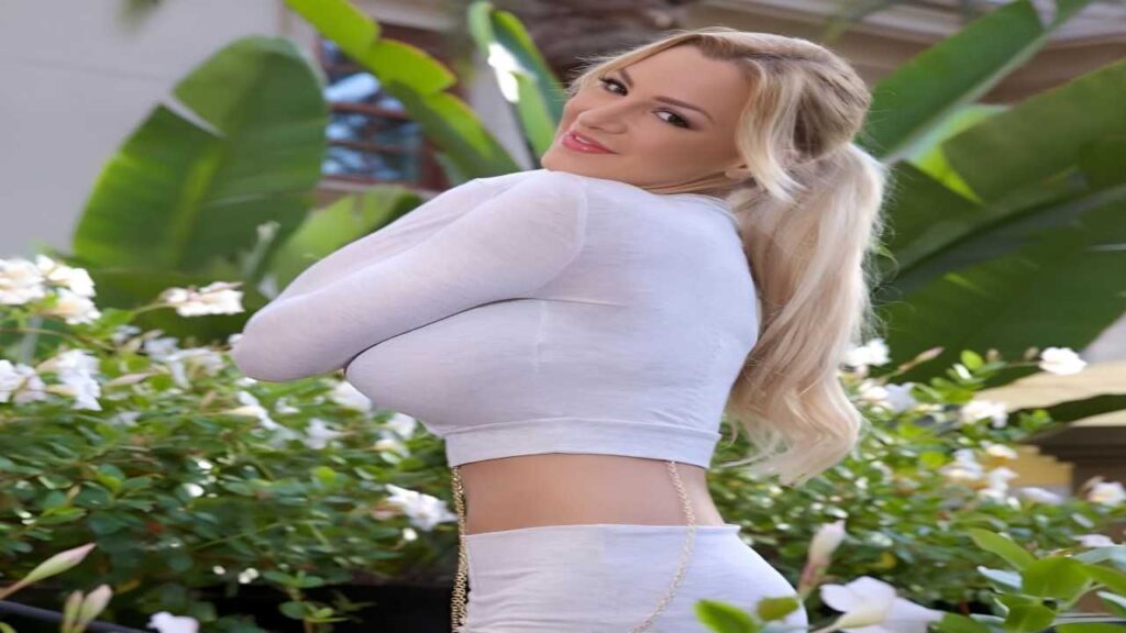 Jordan Carver wearing white pants and a white top stands confidently, showcasing a stylish and modern outfit