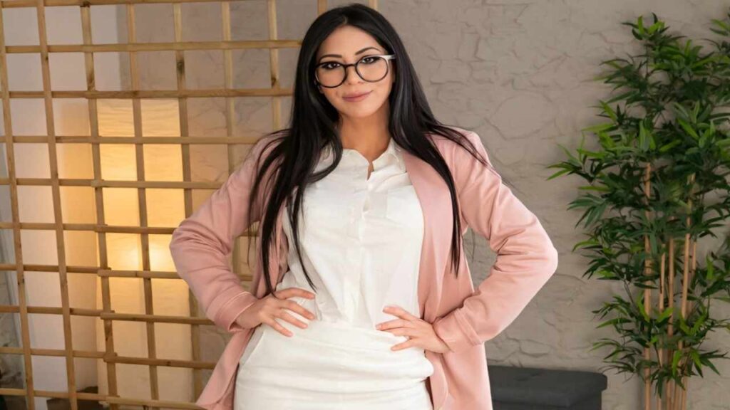 Julia De Lucia wearing glasses and a pink blazer, smiling confidently against a neutral background