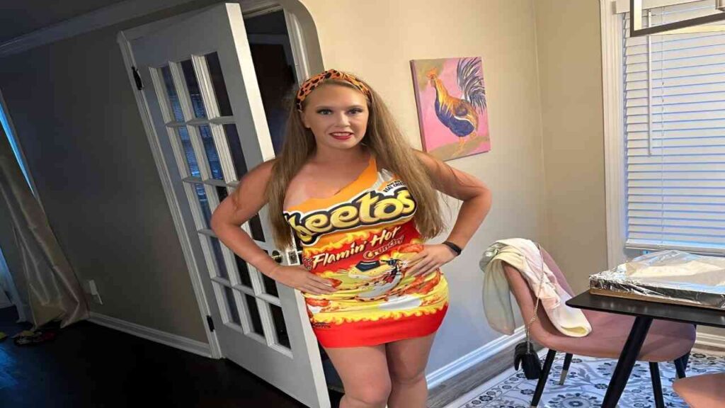 Kagney Linn Karter wearing a dress balances a bag of chips on her head, smiling playfully at the camera