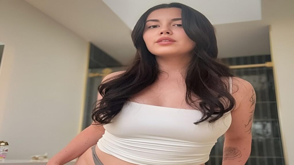 Kayla Summers in a white top and panties stands in a bathroom, looking at herself in the mirror.