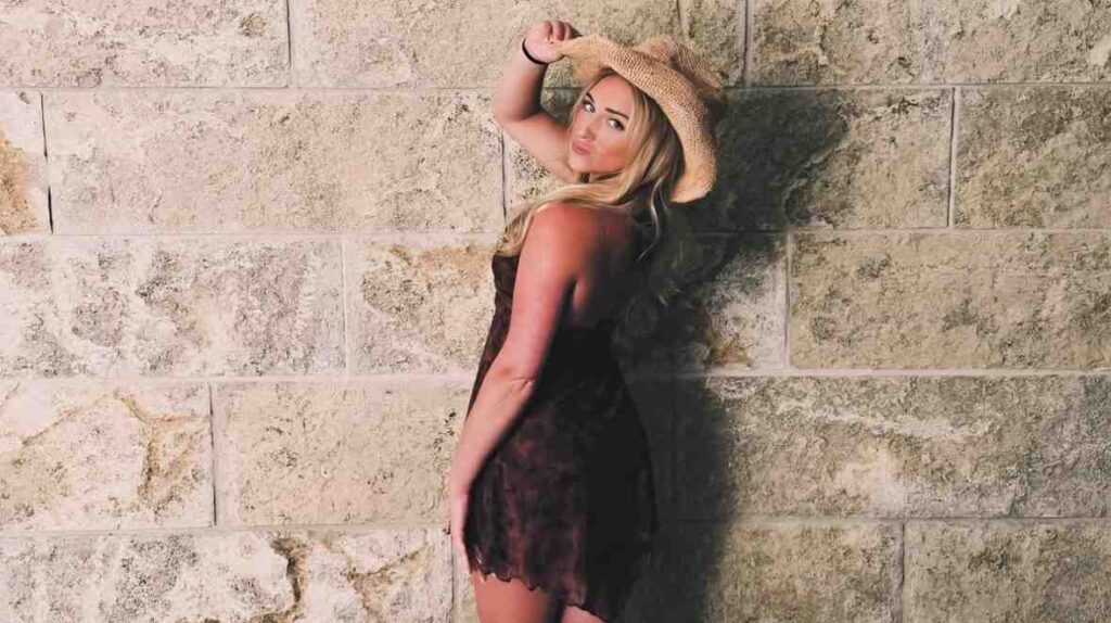 Kelly Madison in a dress and hat leans against a wall, exuding a relaxed and stylish demeanor