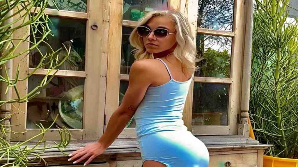 Kelsey Kane in a blue dress leans against a wooden bench, smiling softly in a serene outdoor setting