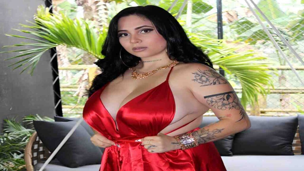 Kim Velez wearing a red dress, showcasing her tattoos, stands confidently with a vibrant expression