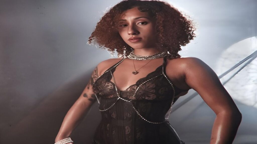 Kira Perez wearing a black lingerie top and a necklace, posing confidently against a neutral background