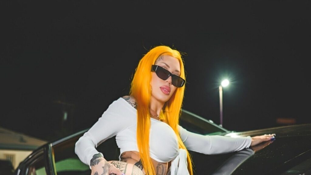 Lacey Jayne with orange hair and tattoos sits casually on a parked car, exuding a relaxed and confident vibe.