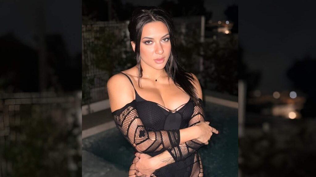 Lena Coxx in a black bodysuit poses confidently beside a sparkling pool under bright sunlight.
