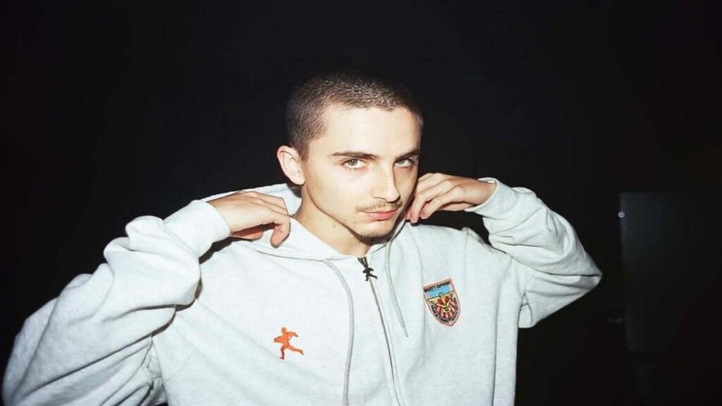 Lil Timmy Tim a hoodie poses for a photo, smiling confidently at the camera.