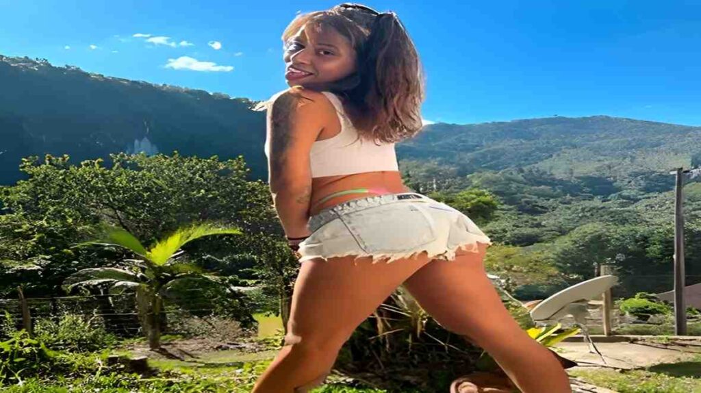 Lolah Vibe in short shorts stands confidently on a hill, overlooking a scenic landscape under a clear blue sky.