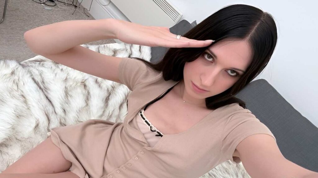 Nbnabunny in a tan dress sits on a bed, looking thoughtfully to the side in a softly lit room.