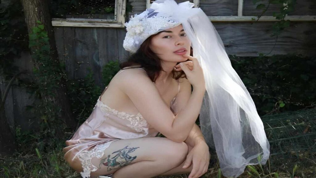 Olive Glass in a wedding dress kneels gracefully, showcasing the intricate details of her gown against a soft background.
