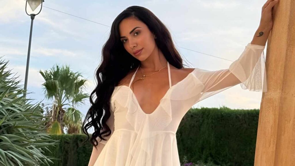 Raquel Alvarez in a white dress poses gracefully on a patio, surrounded by greenery and sunlight.