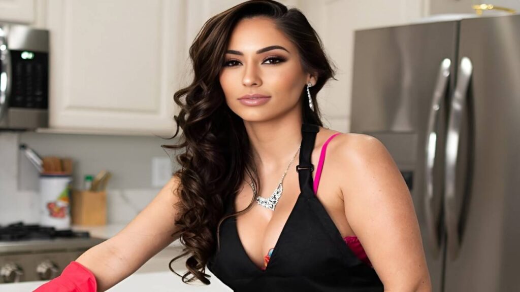 Reena Sky wearing a black apron and pink gloves sits on a kitchen counter, smiling and looking at the camera