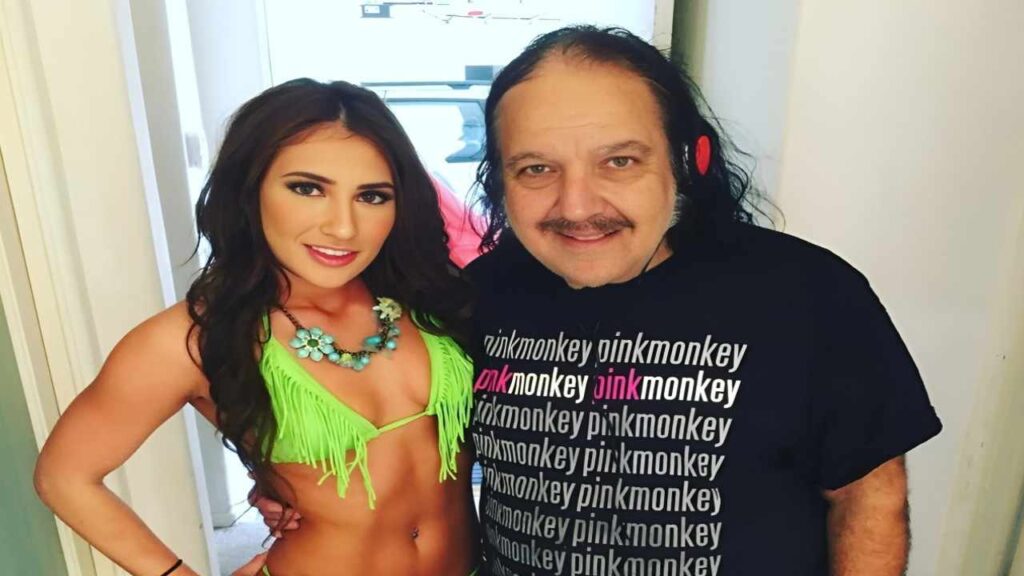 Ron Jeremy wearing a top and a woman in a green bikini pose together, smiling at the camera