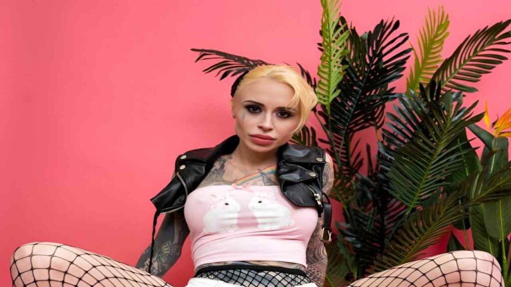 Sasha Beart with tattoos sits against a vibrant pink background, showcasing her unique style and personality.
