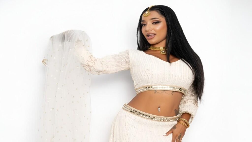 Sasha Prasad in a white outfit poses confidently for the camera, smiling and looking directly at the viewer