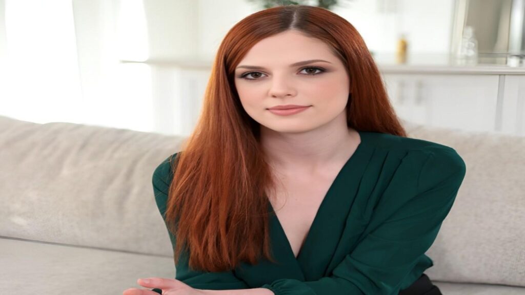 Scarlett Mae with red hair is sitting comfortably on a couch, looking relaxed and engaged in her surroundings