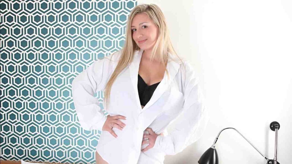 September Carrino in a white robe stands on a bed, looking serene and relaxed in a softly lit room.