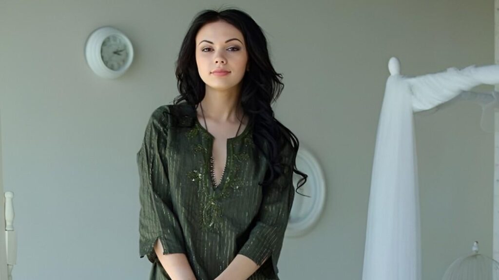 Sheri Vi in a green shirt stands in a cozy bedroom, surrounded by soft lighting and neutral-colored decor
