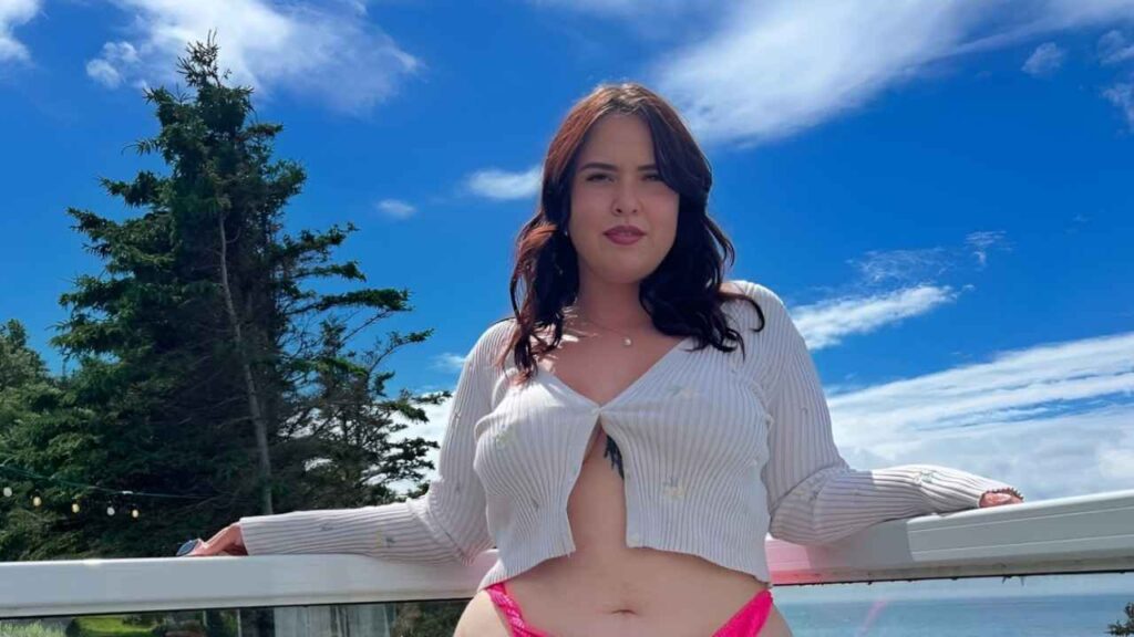 Steph Oshiri wearing a pink bikini and a white sweater stands confidently, showcasing a relaxed beach style