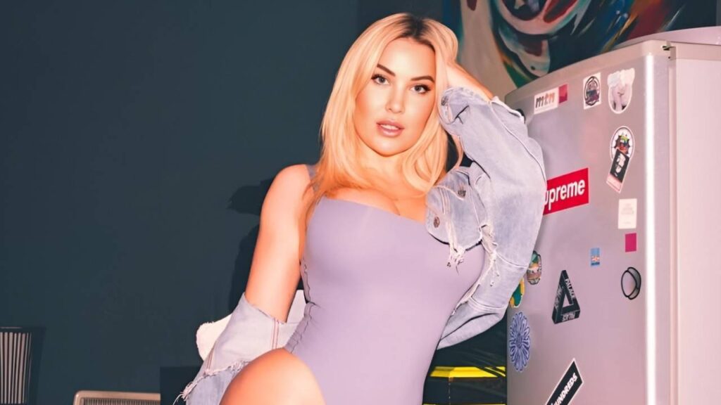 Amber Jade in a purple bodysuit poses confidently next to a modern refrigerator in a bright kitchen setting