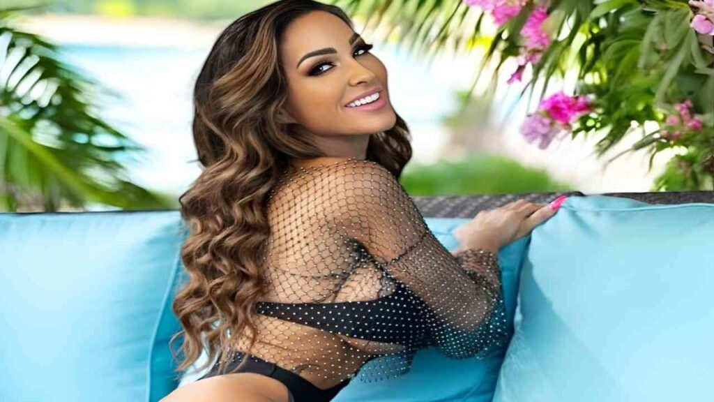 Aubrey Black wearing a black fishnet top sits on a blue couch, exuding a relaxed and stylish vibe.