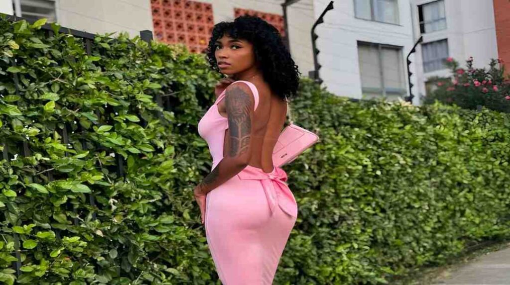 Barbie Rous in a pink dress stands on the sidewalk, smiling and looking towards the camera.