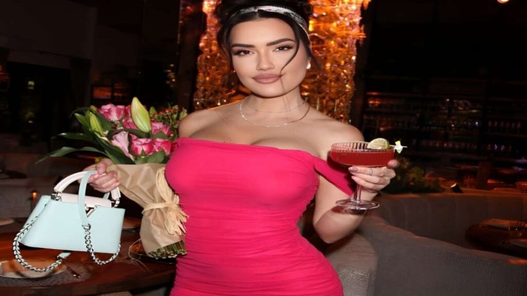 CJ Sparxx in a pink dress holds a drink in one hand and a bouquet of flowers in the other, smiling brightly