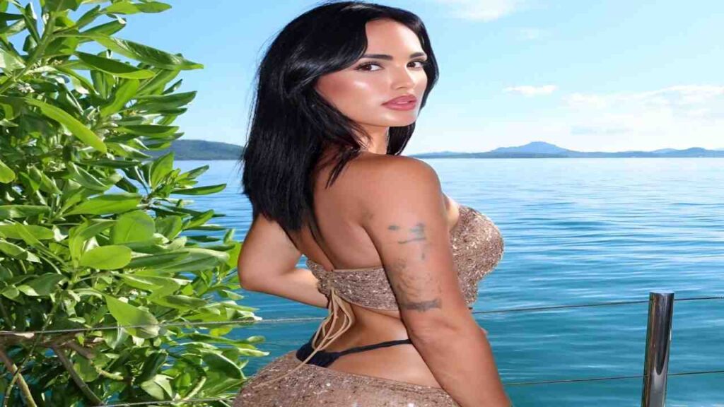 Claudia Bavel in a bikini poses confidently by the water, showcasing a sunny beach setting.